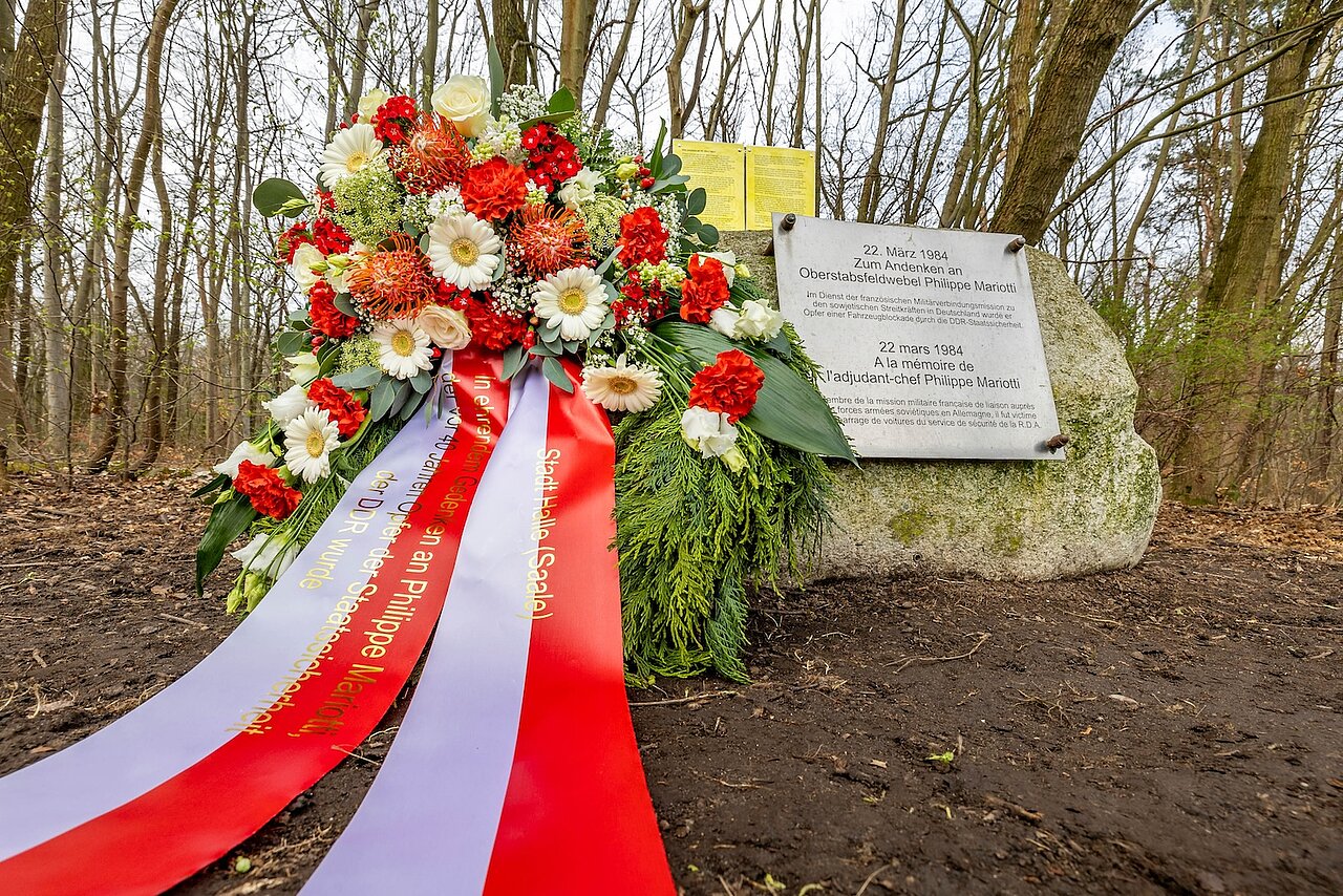 Vor dem Gedenkstein im Wald liegt ein Kranz mit roten und weißen Blumen. Auf der Banderole steht Stadt Halle (Saale)