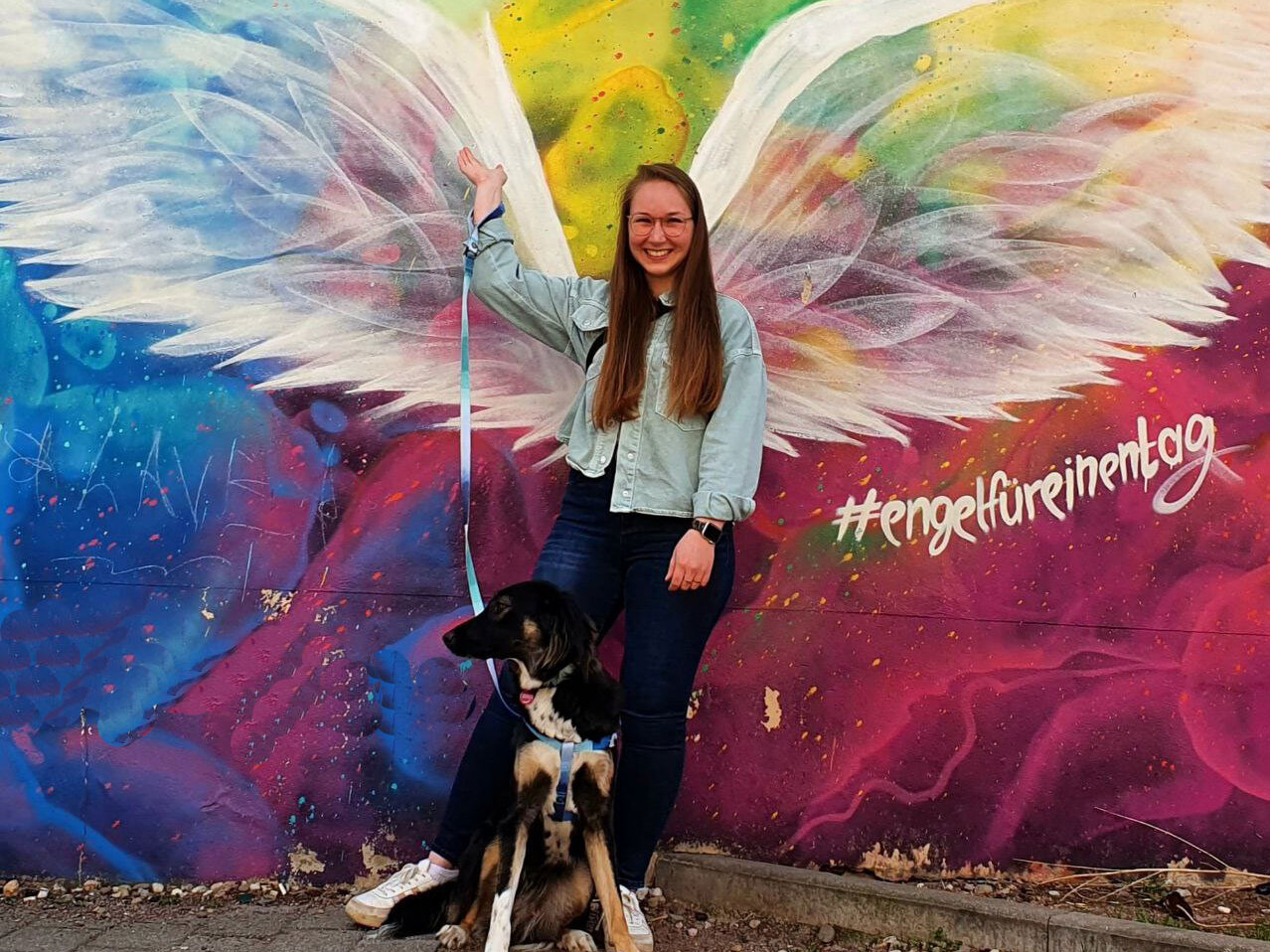 eine junge Frau steht mit ihrem Hund vor einer mit Engelsflügeln besprühten Hauswand